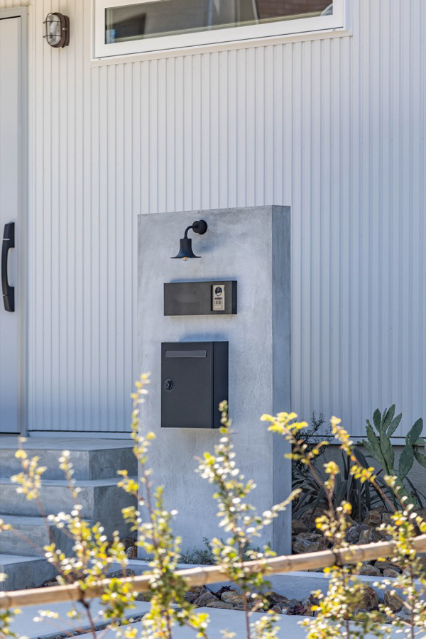 白いガルバリウムとモルタルで仕上げた門柱にドアフォン、ポスト、照明器具がついている。